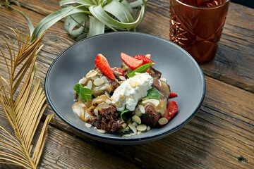 Appetizing brownie dessert cake with ice cream, strawberries and almonds in a gray bowl on a wooden background