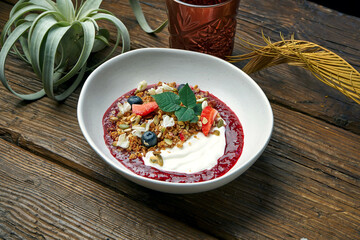 Sweet and healthy breakfast - granola with yogurt, raspberry jam and summer fruits in a bowl on a wooden background