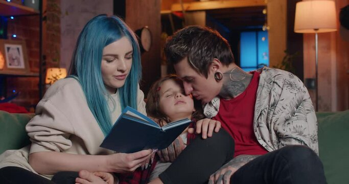 Portrait of young modern family spending time together with book in evening. Beautiful mom reading aloud while her husband with tattoos and tunnels listening and kissing their sleeping daughter.