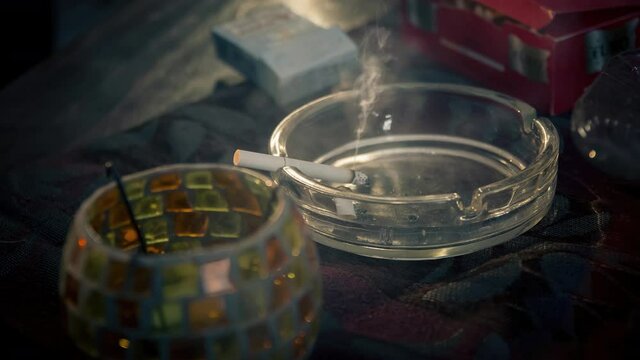 Cinemagraph of smoke emitted from a burnt cigarette in a glass ashtray. It placed on a tablecloth on a wooden table. Close up, loop with selective focus.