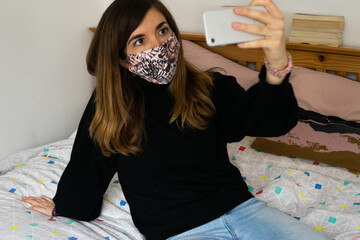 Photo of a young and attractive woman with a black jumper wearing a reusable face mask taking a selfie with her phone sitting on the bed