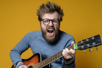 Funny hipster in an old sweater plays an acoustic guitar and sings loudly, on a yellow background. Hobbies, lifestyle.