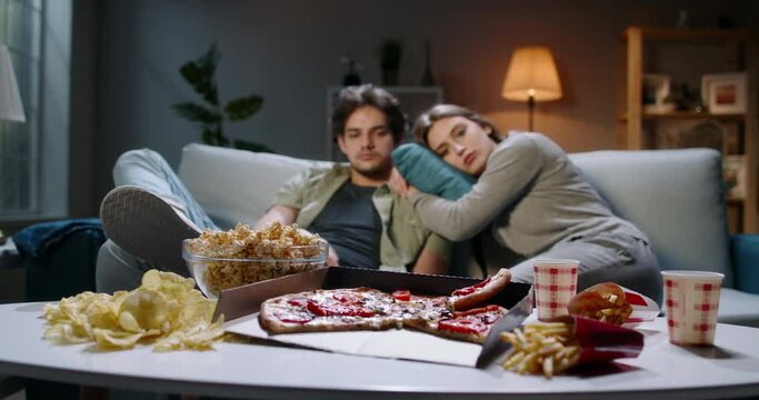 Young caucasian couple extremely frustrated. Bored students are sitting in front of tv, switching channels with remote and eating unhealthy food, tired after hard day 4k footage