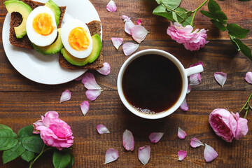 Tasty breakfast with avocado toasts, coffee, and boiled eggs on a wooden background with beautiful roses and rose petals.  Breakfast concept. Top view, flat lay, layout
