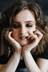 Fototapeta premium Close-up portrait of a beautiful girl with closed eyes with professional makeup on a dark background who holds her hands to her head. Photography of a cute girl