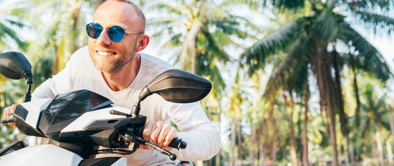 Happy smiling male tourist in sunglasses riding motorbike scooter during his tropical vacation under palm trees. © Soloviova Liudmyla