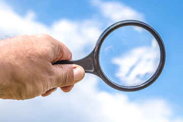 Man's hand holding a magnifier or magnifying glass with blue sky and white cloud background. Free space for advertisement text.
