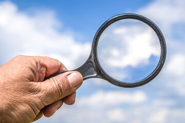 Man's hand holding a magnifier or magnifying glass with blue sky and white cloud background. Free...
