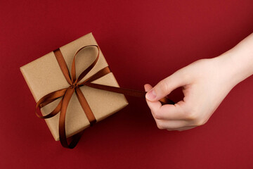 Female hands hold a gift box wrapped in craft paper and tied with a brown ribbon on a red background close-up.