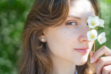 Portrait of a young woman close-up, covering her eye with a branch of jasmine. Beautiful smiling girl with flower in her hand
