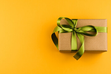 A gift box wrapped in craft paper and tied with a green ribbon on a yellow background close-up. Copy space