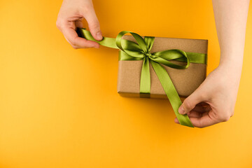 Female hands hold a gift box wrapped in craft paper and tied with a green ribbon on a yellow background close-up. Copy space