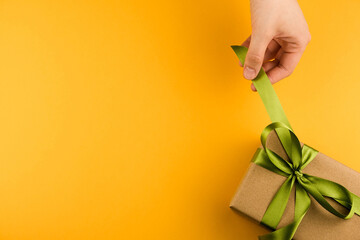 Female hands hold a gift box wrapped in craft paper and tied with a green ribbon on a yellow background close-up. Copy space