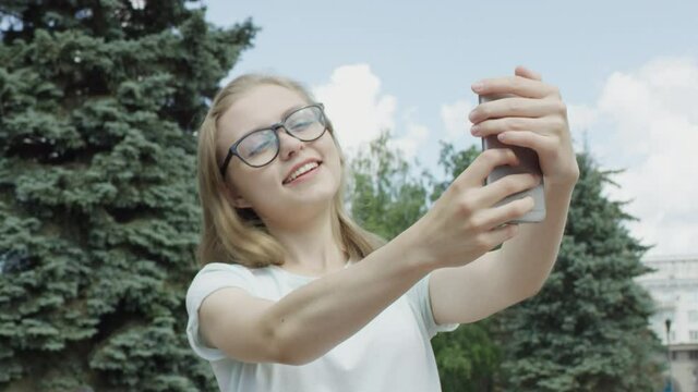 Pretty blonde girl in glasses posing and smiling for selfie against fir tree and blue sky in park on sunny summer day. Camera floating around.