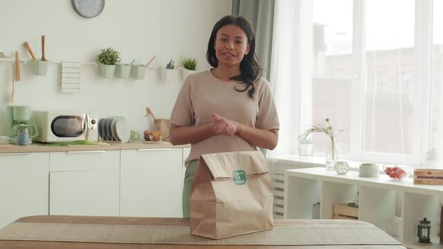 Medium shot of young mixed-race woman with dark hair is standing at kitchen table and unpacking delivered package with cooked food while commenting her actions on camera