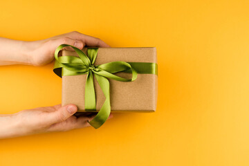 Female hands hold a gift box wrapped in craft paper and tied with a green ribbon on a yellow background close-up. Copy space