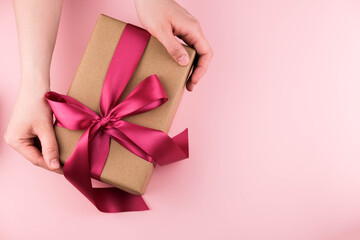 Female hands hold a gift box wrapped in craft paper and tied with a pink ribbon on a pink background close-up. Copy space