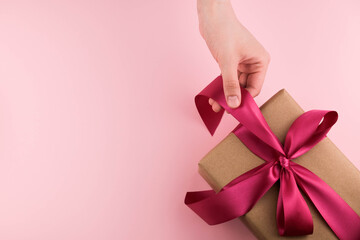 Female hands hold a gift box wrapped in craft paper and tied with a pink ribbon on a pink background close-up. Copy space