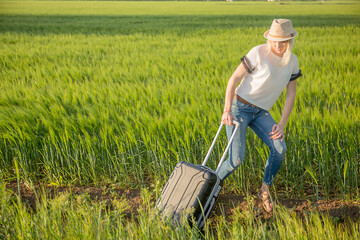 Sexy blonde girl in a hat with a suitcase on a background of green field, with space for text