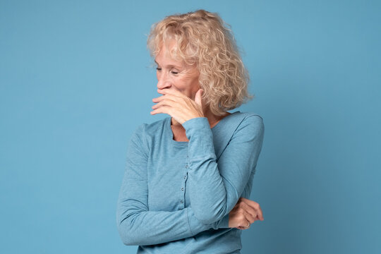 shy caucasian mature woman smiling with hand on her mouth. Studio shot on blue wall.