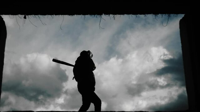 A guy in a hard hat waves a baseball bat against a dark sky. Around the destroyed building