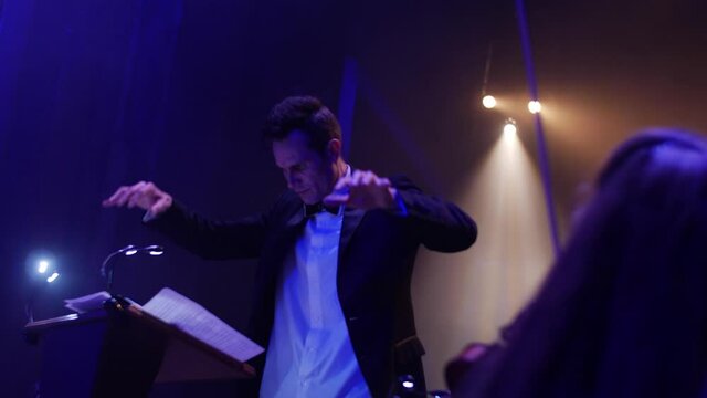 male conductor stands in blue light waves his hands for an orchestra at a concert