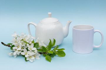 White cup and kettle without logos on a blue background. Nearby lies a branch of a flowering apple tree. The concept is a layout for advertising. Tea party. Photo in pastel tones