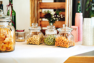 Salty bar of several kinds of nuts and chips in glass jars decorated on wooden table with white table cloth. Wedding or party concept.