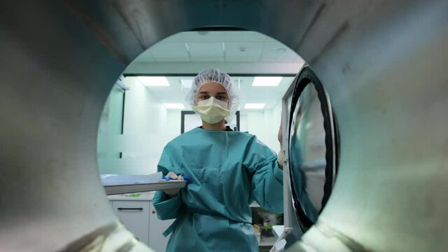 Laboratory assistant in green uniform, yellow mask and white cap takes out packaging with dental tools from autoclave sterilizer machine after disinfection. POV medical oven. Modern professional