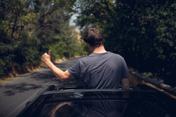 Young happy man drives a car and holds his hand out from the window. Driver enjoys driving and shows Ok sign with his hand out of window. Road trip, travel and freedom concept.