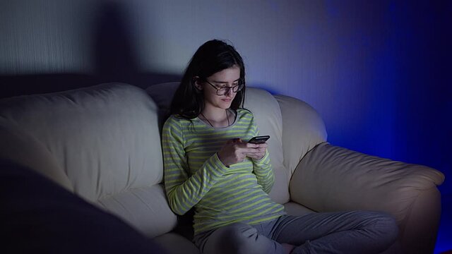 Girl in glasses sits on the couch next to the TV with her legs crossed and reads correspondence in her phone