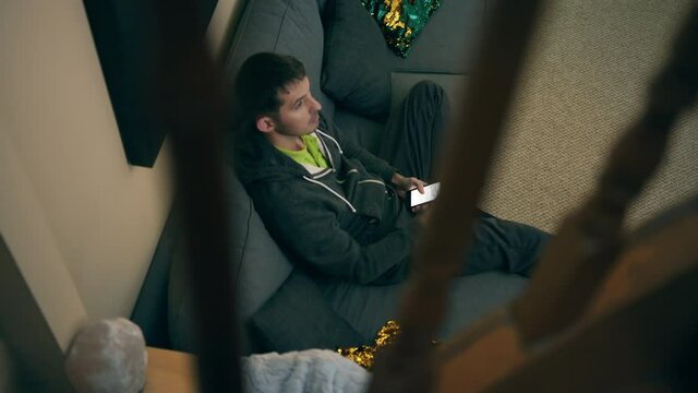 High quality close-up 4k slow motion video of a man sitting on a couch in his detached beautiful house main floor and watching television while browsing on the phone.