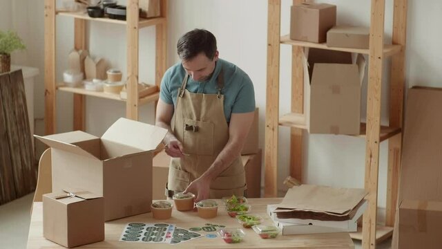 Medium shot of Caucasian man wearing apron standing at wooden table in office and packing box for delivery