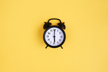 View from above on black alarm clock on yellow background. 6 a.m. Time is now