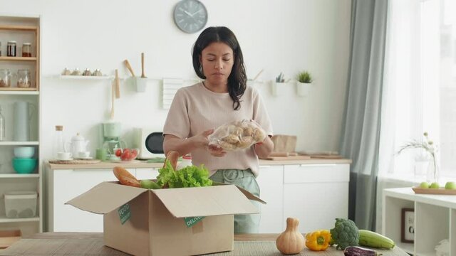 Medium shot of young mixed-race woman unpacking delivered box with food and commenting her actions on camera