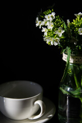 wild white medium-sized flowers in a glass jar with a white tea pair on a dark background in the sunlight