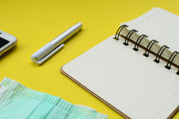 a blank notebook with a face mask, and a pen on yellow background.