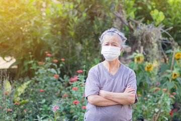 A portrait of an elderly woman wearing a face mask and arms crossed while standing in a garden