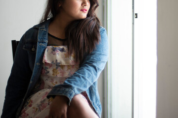 Indian Bengali beautiful and cute brunette girl in a casual blue jeans shirt and white top sitting on a chair inside a room facing a glass door with sidelight. Indian lifestyle and fashion portrait