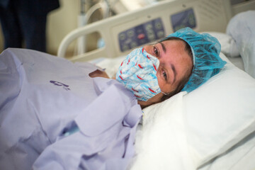 Mother in labor pains with tears in her eyes waiting on child to be born in hospital wearing mask gown and hair net in hospital bed waiting for cesarean section C-section child birth during Covid-19