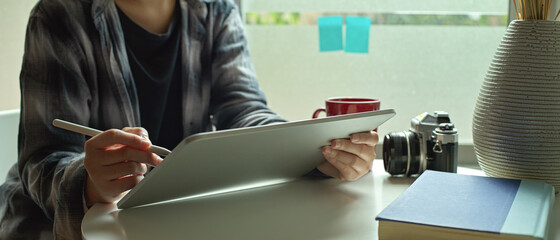 Female freelancer working on digital tablet with stylus pen on coffee table beside window