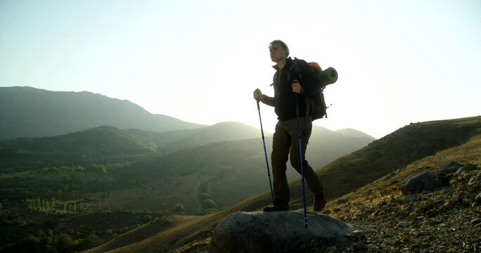 Young caucasian adventurer hiking in mountains during sunset, then stopping to look in distance - active lifestyle, nature, travel, freedom concept 4k footage