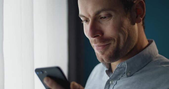 Close up of happy young guy in trendy shirt using smartphone while standing near window at home. Concept of modern technology and free time.