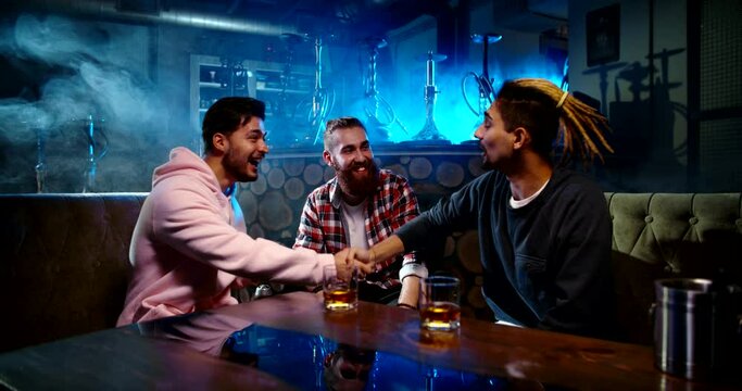 Three young male friends relaxing in hookah bar, smoking a shisha and drinking whisky, throwing a bachelor party - relaxation, lifestyle, recreational pursuit concept 4k footage