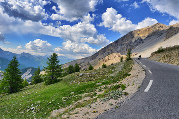 Motorbike rider in the distance rear view on the ride in mountain landscape under cloudy blue sky Italy Piedmont Alps circa June 2015.