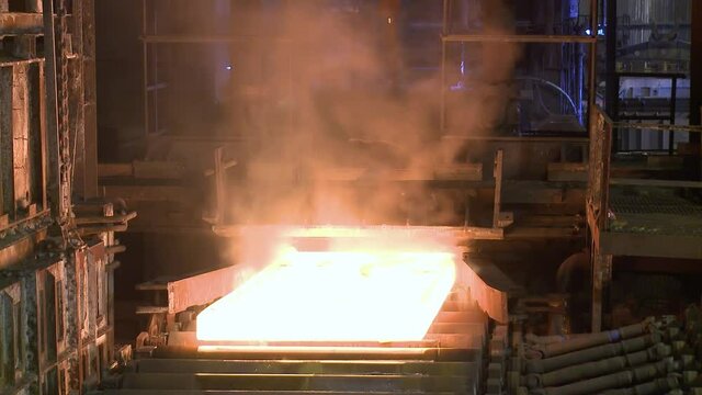 White-hot rectangular steel slab moves on rollers to cool at steel production plant.