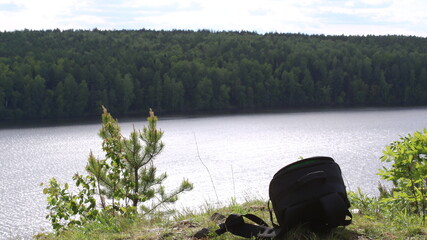 Obraz premium A moment from a trip by a mountain lake with a black backpack left on a cliff of a cliff against the background of a smooth surface of water and a dark forest on the other side