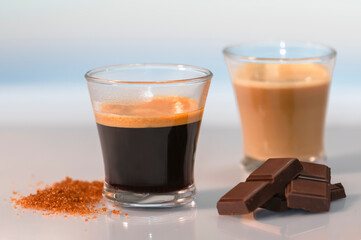 an espresso coffee surrounded by brown sugar, chocolate squares and an out of focus cortado at the back on a glass table