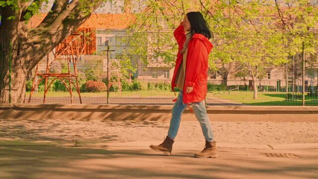 side view woman with fluttering long hair in casual shoes and red raincoat walking in residential area slow motion on the background playground spring season in sunlight