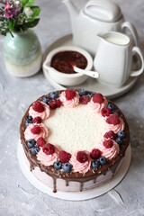 chocolate sponge creamy cake with berries and coconut flakes on grey table with strawberry jam and white retro tea set